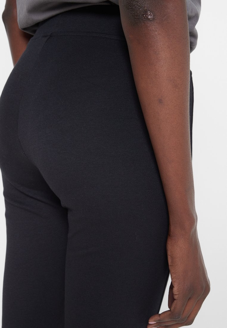 Persona vistiendo pantalones ajustados negros y una camiseta gris, mostrando el lado derecho desde la cintura hasta la mano contra un fondo blanco.