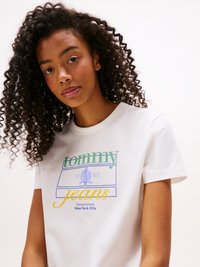 Jeune femme aux cheveux bouclés portant un T-shirt blanc avec le texte "tommy jeans" et "Established New York City 1985" en vert, bleu et jaune.