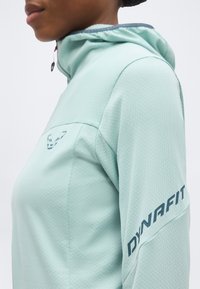 Άτομο που φοράει ανοιχτό μπλε μπουφάν με κουκούλα με την επιγραφή «DYNAFIT» στο μανίκι και ένα μικρό λογότυπο αιλουροειδούς στο στήθος, εμφανίζεται από το πλάι.