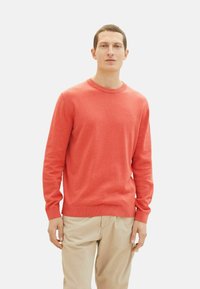 Koral pullovertrøje, rund halsudskæring, ribbede manchetter og kant, letvægtsstof, bæres med beige bukser, model står mod en hvid baggrund.