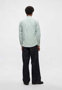 Ανοιχτό πράσινο πουκάμισο με μακριά μανίκια, διαθέτει πίσω ωμοπλάτη, μανσέτες με κουμπιά και άνετη εφαρμογή, συνδυασμένο με σκούρο μπλε τζιν παντελόνι με φαρδιά μπατζάκια και μαύρα παπούτσια.