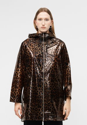 Nő fényes barna és fekete leopárdmintás kapucnis esőkabátban, elülső cipzárral, egyszínű világos háttér előtt áll.