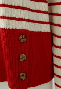 Pull tricoté rouge et crème avec des rayures rouges horizontales et trois boutons foncés alignés verticalement sur la section rouge.