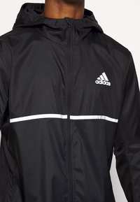 Svart Adidas-hoodie med framdragkedja, vit horisontell rand över bröstet och Adidas-logga på vänster sida.