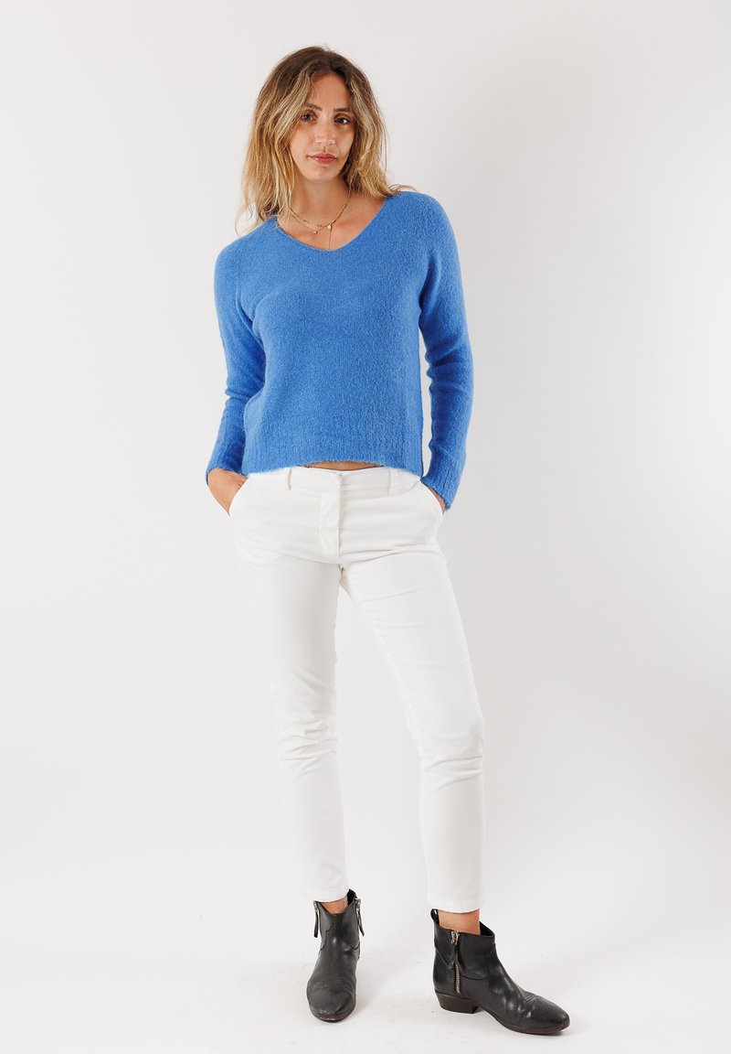 Maglione azzurro lavorato a maglia con scollo a V, maniche lunghe e finitura testurizzata, abbinato a pantaloni bianchi a vestibilità aderente e stivaletti neri alla caviglia.