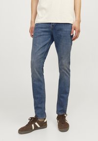 Jeans de mezclilla azul con un corte ajustado, con un ligero desvanecimiento, cinco bolsillos y una cintura de altura media, combinados con zapatillas marrones.