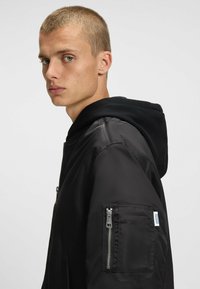 Veste bomber noire avec une poche zippée et une capuche noire. Matière lisse et brillante, offrant une coupe ajustée et un détail de marque subtil sur la manche.