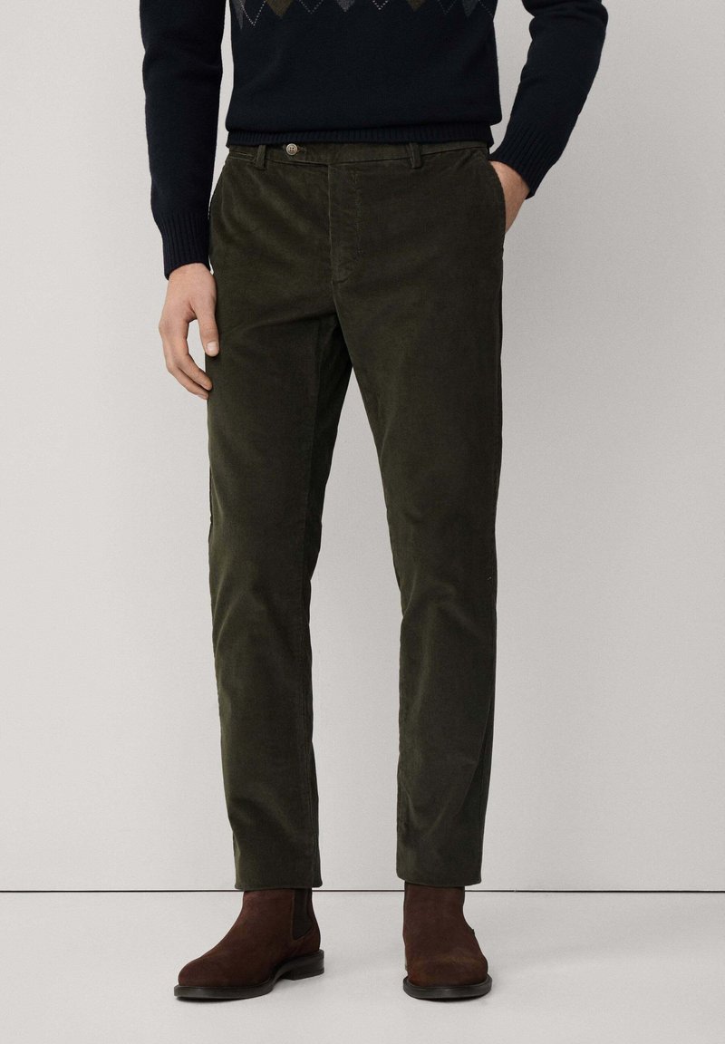 Pantaloni di velluto a coste verde scuro con vestibilità su misura, dotati di zip, tasche frontali e una texture sottile, abbinati a stivali alla caviglia marroni.