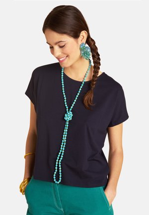 Lächelnde Frau mit geflochtenen Haaren trägt ein schwarzes T-Shirt, eine türkisfarbene Perlenkette, große blau-grüne Ohrringe, türkise Hosen und goldene Armreifen.