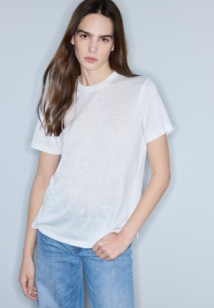 Junge Frau mit langen braunen Haaren, die ein schlichtes weißes T-Shirt und blaue Jeans trägt, steht vor einem hellgrauen Hintergrund.