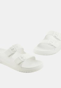 Chanclas blancas sin hebillas, hechas de goma, con dos correas ajustables que cuentan con hebillas, una suela texturizada y un diseño minimalista.