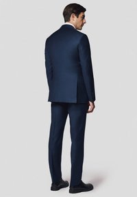Granatowy garnitur z dopasowaną marynarką i prostymi spodniami, wykonany z gładkiej tkaniny. Tylny widok pokazuje pojedyncze rozcięcie i eleganckie buty.