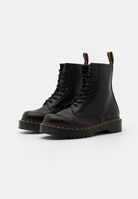 Bottes de cheville en cuir noir avec un bout rond, un design à lacets, des coutures jaunes et une semelle en caoutchouc texturée. Dotées d'une languette pour un enfilage facile.