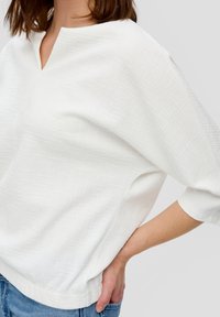Weißes, strukturiertes Oberteil mit einem dezenten Karomuster, V-Ausschnitt und Dreiviertelärmeln. Weicher Stoff mit lässiger Passform.
