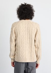 Béžna pletena jopica z teksturiranim vzorcem, rebrastimi manšetami in robom ter sproščenim krojem. Preja vsebuje bleščice barv skozi celotno površino.