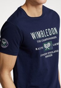Muž v námořnickém modrém tričku s logem Wimbledonu, zkříženými tenisovými raketami a textem odkazujícím na šampionát a Ralph Lauren.
