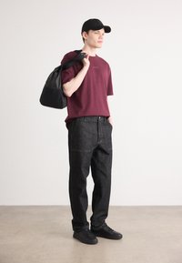 Bordeauxfarbene Kurzarm-T-Shirt mit Logo, schwarze High-Waist-Jeans und schwarze Sneakers. Trägt eine schwarze Umhängetasche. Neutrale Hintergrund.
