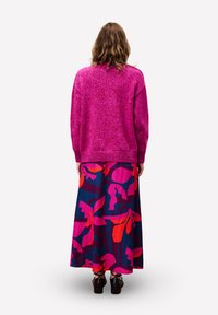 Personne ayant des cheveux ondulés, portant un pull rose vif et une longue jupe avec de grands motifs floraux roses et oranges, se tenant de dos.