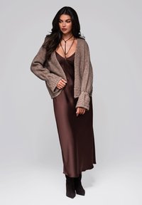 Braune Strickjacke über einem braunen Satinkleid, kombiniert mit kniehohen schwarzen Stiefeln und gestapeltem Schmuck. Weiche Texturen und lässige Passform.