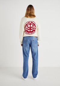 Crème gebreide trui met een rood cirkelontwerp op de achterkant, gecombineerd met lichtblauwe rechte spijkerbroek en witte sneakers.