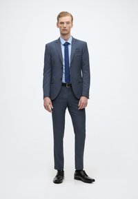 Fato cinza com uma textura suave, calças a condizer e uma camisa azul clara. O conjunto apresenta uma gravata azul-marinho e sapatos de vestir pretos.