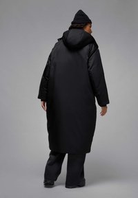 Cappotto lungo nero in piuma con cappuccio, caratterizzato da una texture liscia e una vestibilità oversize. Abbinato a pantaloni scuri e un berretto nero lavorato a maglia.