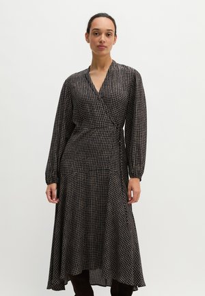 Femme portant une robe portefeuille manches longues avec un col en V et un motif à carreaux noir et beige, debout devant un fond clair uni.