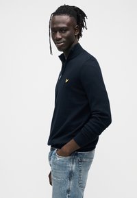 Laivastonsininen puolivetoketjullinen neule, joka on valmistettu pehmeästä neuloskankaasta ja jossa on keltainen kirjailtu logo rinnassa, yhdistetty vaaleansinisillä rikotuilla farkuilla.