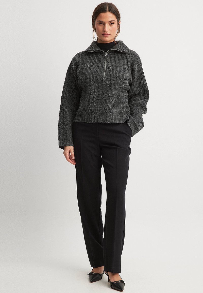 Sivý rebrovaný pulóver s polovičným zipsom a golierom, spárovaný s upravenými čiernymi nohavicami. Má uvoľnený strih a dlhé rukávy; hladký povrch.