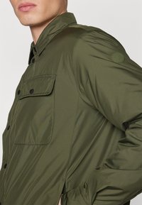 Giacca verde oliva con colletto bottonato, dotata di taschino e un patch del logo sulla manica. Realizzata in un materiale liscio e leggero.