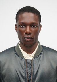 Jaqueta bomber em tecido cinza com uma textura suave, gola canelada verde e fecho de zíper frontal. Camisa de cor clara com colarinho visível por baixo.