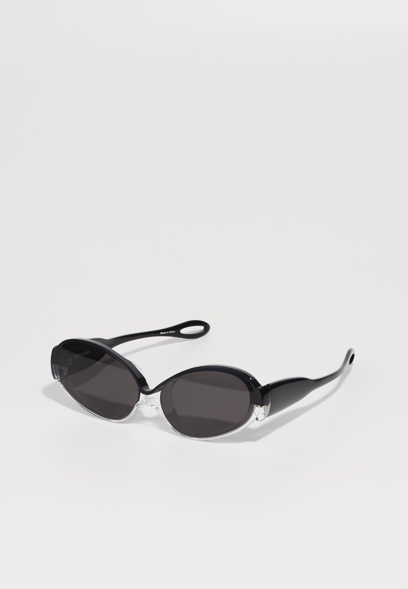 Melni un caurspīdīgi ovāli saulesbrilles ar tumšiem stikliem un elegantiem, izstieptiem kātiem. Rāmim ir spīdīga apdare. Ievērojami caurspīdīgi akcenti priekšpusē.