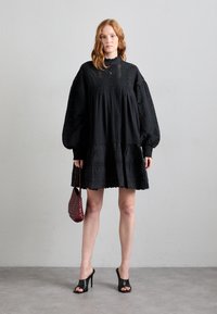 Črna oversize obleka z dolgimi balonastimi rokavi, teksturirano blago, volanast rob in podrobnost z gumbi spredaj. V kombinaciji s temno rdečo torbico in črnimi petami.