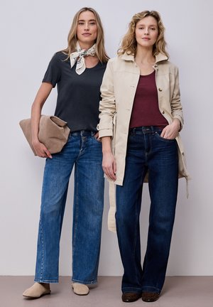 Twee vrouwen staan naast elkaar, gekleed in casual outfits met jeans; de ene draagt een zwart T-shirt en sjaal en houdt een beige clutch vast, de ander een beige jas.