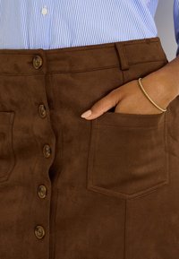 Falda de ante marrón con cierre frontal de botones, dos bolsillos laterales y textura suave, combinada con una camisa azul de rayas encima.