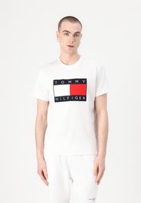 Valkoinen puuvillapaita, jossa on suorakulmainen logo, jossa on punaisia, valkoisia ja tummansinisä alueita sekä teksti "TOMMY HILFIGER" paksuilla kirjaimilla.
