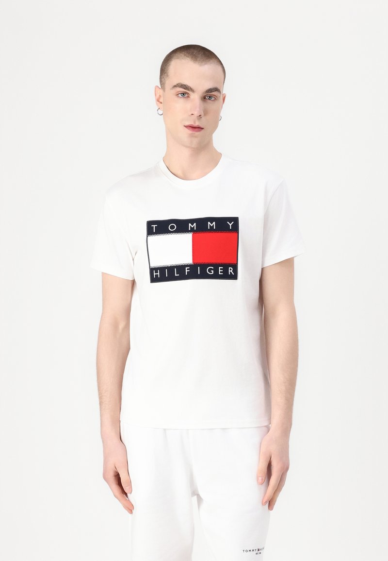 Valkoinen puuvillapaita, jossa on suorakulmainen logo, jossa on punaisia, valkoisia ja tummansinisä alueita sekä teksti "TOMMY HILFIGER" paksuilla kirjaimilla.