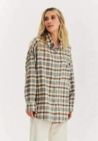 Kobieta o długich blond włosach, ubrana w dużą koszulę w beżowo-szaro-brązową kratę na guziki oraz białe spodnie, stojąca na jednolitym tle.