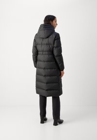 Schwarze Daunenjacke mit Kapuze, mit horizontalen gesteppten Abschnitten, die bis zur Mitte der Wade reicht. Getragen über dunklen Jeans und schwarzen Schuhen.