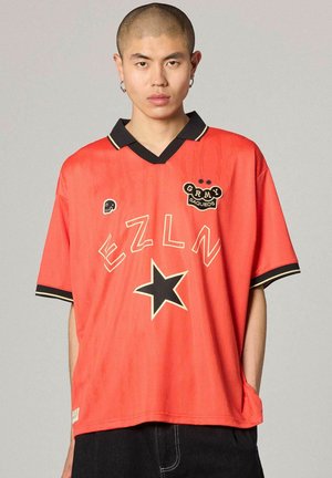 Giovane con capelli corti che indossa una camicia rossa oversize con stella nera, testo "EZLN" e toppa "GRMY SAQUEOS", mani nelle tasche.