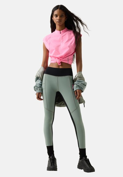 Kvinde iført grønne leggings, pink ærmeløs lynlåsbluse knyttet om livet, sorte sneakers, holder en lysegrøn jakke bag ryggen.