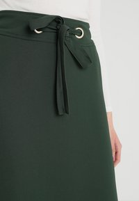 Jupe verte en forme d'aile de mouette, fabriquée en tissu lisse, dotée d'une ceinture à nouer avec des œillets en métal. Le design inclut une taille nette et une coupe fluide.