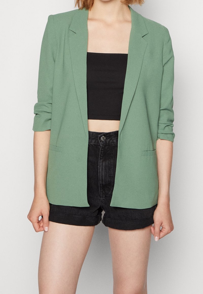 Personne portant un blazer vert clair sur un haut court noir et un short noir taille haute sur un fond uni.