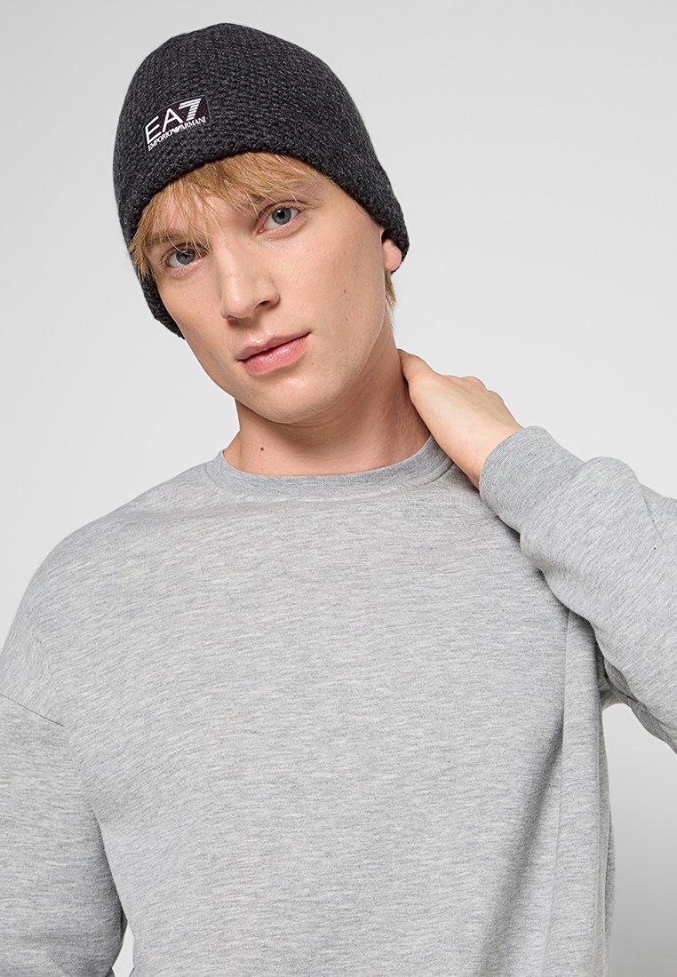 Szara bluza z okrągłym dekoltem; czarna wełniana czapka z białym logo. Model wydaje się patrzeć bezpośrednio w obiektyw.