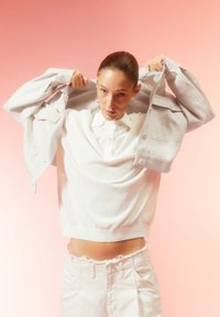 Weißer Pullover mit strukturiertem Finish, kombiniert mit hellen Hosen. Das Modell zieht eine hellgraue Jacke über die Schultern.