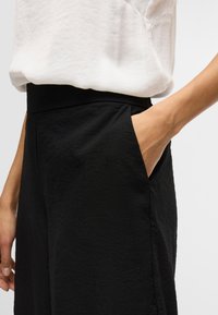 Pantalones negros con una tela texturizada, que cuentan con una cintura alta y bolsillos laterales. Combinados con una blusa blanca de corte holgado.