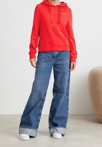 Sudadera roja con cordón, combinada con vaqueros anchos azules. Zapatillas blancas contrastan con el conjunto. Las texturas son suaves y casuales.