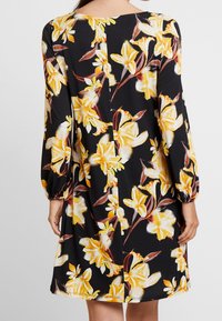 Vestido floral negro con mangas largas abullonadas, que presenta flores amarillas y blancas. Tejido suave, a la altura de la rodilla con un escote redondeado y costura en la parte trasera.