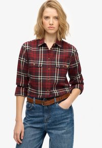 Camisa de franela a cuadros rojos y marinos con dos bolsillos en el pecho, cuello abotonado y mangas enrolladas, combinada con pantalones de mezclilla azules y un cinturón marrón.