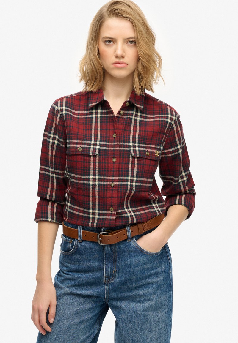Camisa de franela a cuadros rojos y marinos con dos bolsillos en el pecho, cuello abotonado y mangas enrolladas, combinada con pantalones de mezclilla azules y un cinturón marrón.